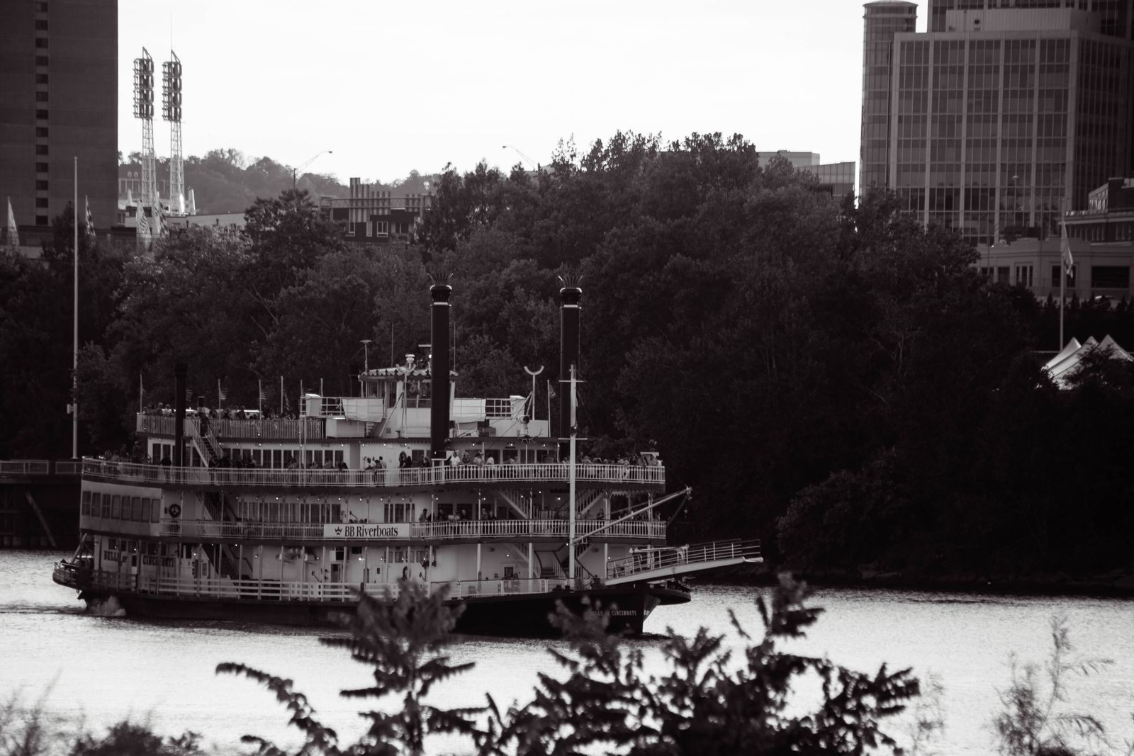 Black-and-white photo of a twin-stacked sternwheel riverboat passing city buildings, like a steamboat cruise in New Orleans